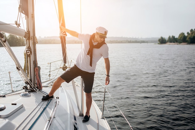 handsome and serious sailor standing on his yacht. He holds on tube and look at edge of boat. Young man poses.