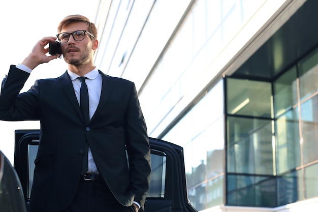 Красивый серьезный молодой бизнесмен, стоящий возле своего нового комфортабельного автомобиля и идущий в офис.