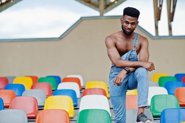 Handsome sexy african american bare torso man at jeans overalls posed on colored chairs at stadium Fashionable black man portrait