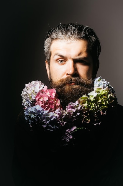 Handsome sexy bearded young man hipster with floral beard and mustache on serious face in studio holding hydrangea flower bouquet on grey background closeup