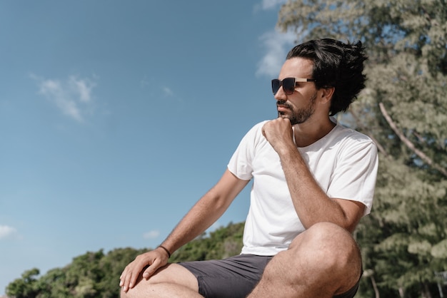 Handsome sexy latin brunette man with a beard in sunglasses and a white T-shirt in profile against a tropical landscape