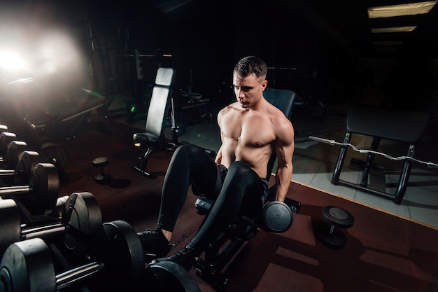 Handsome shirtless guy lifting heavy dumbbells while sitting on bench in dark gym