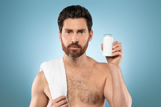 Красивый мужчина без рубашки с полотенцем на плече, показывающий дезодорант, позирующий после душа на синем фоне студии