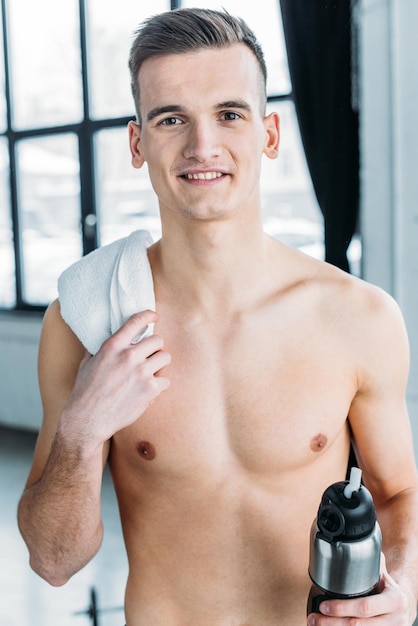 Handsome shirtless sportsman holding sports bottle and smiling at camera in gym