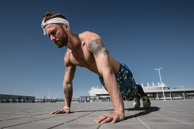 Photo handsome shirtless sportsman looking away doing push up exercise on workout ground outdoors at bright sunny day. sport lifestyle .