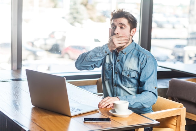 Handsome shocked scared man freelancer working on laptop looking at screen covering mouth with palm