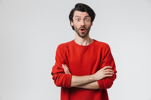 Handsome shocked young bearded brunette man wearing sweater standing isolated over white wall, arms folded