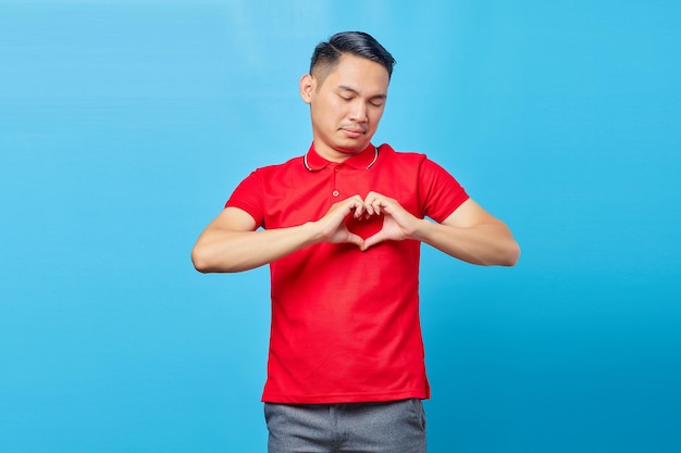 Handsome smiling Asian man in red shirt showing heart gesture isolated over blue background