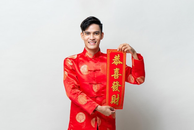 中国の旧正月の概念のための孤立したスタジオライトグレーの背景に赤いスクロールを示すハンサムな笑顔のアジア人、外国のテキストはあなたがより多くの富を得ることができることを意味します