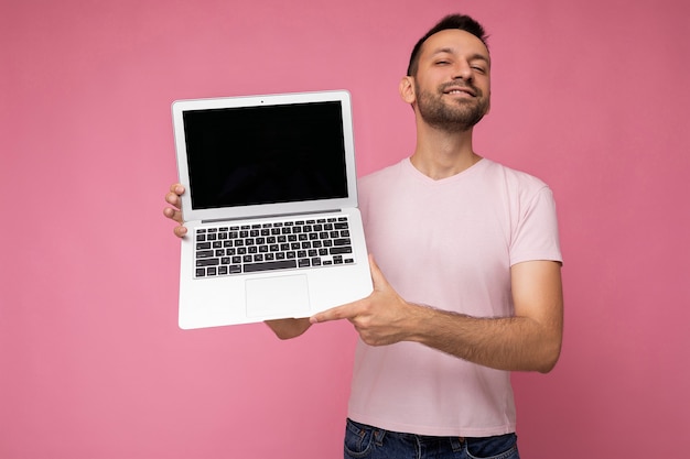 Handsome smiling brunet man holding laptop computer looking at camera in t-shirt on isolated pink background.