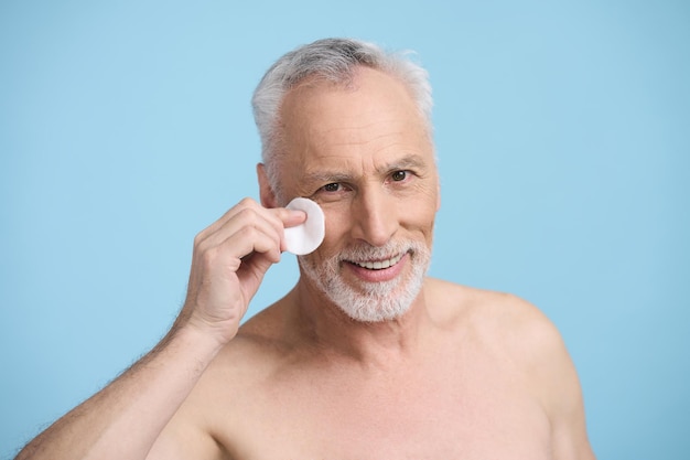 Handsome smiling Caucasian senior man holding cotton pad at his face isolated on blue background