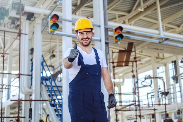 Handsome smiling caucasian worker in overalls and with helmet on head showing thumbs up while standing at refinery.