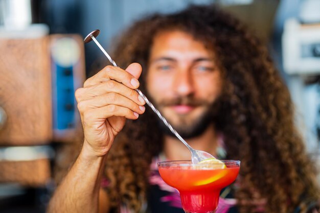 Красивый улыбающийся бармен с кудрявыми волосами готовит коктейль в баре курорта