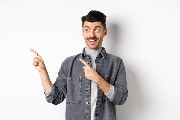 Handsome smiling guy checking out advertisement, looking and pointing left with excited face, standing on white background.