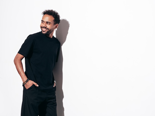 Handsome smiling hipster model Sexy unshaven man dressed in black summer tshirt and jeans clothes Fashion male posing near white wall in studio at sunny day Isolated