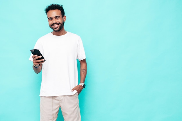 Handsome smiling hipster model Sexy unshaven man dressed in white summer tshirt and jeans clothes Fashion male posing near blue wall Holding smartphone Looking at cellphone screen Using apps