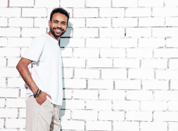 Handsome smiling hipster model Sexy unshaven man dressed in white summer tshirt and jeans clothes Fashion male posing near white brick wall in studio at sunny day Isolated