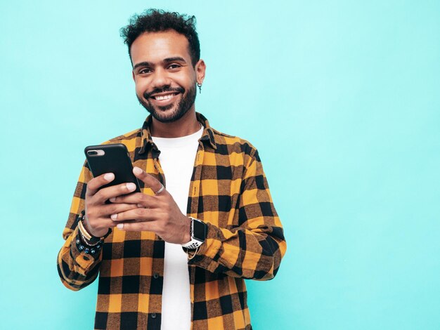 Handsome smiling hipster model Sexy unshaven man dressed in yellow summer shirt and jeans clothes Fashion male posing near blue wall Holding smartphone Looking at cellphone screen Using apps