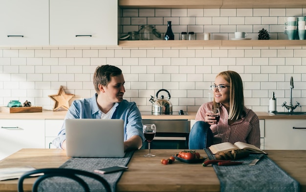 Handsome smiling man sitting and working with laptop while looking at young girlfriend in glasses