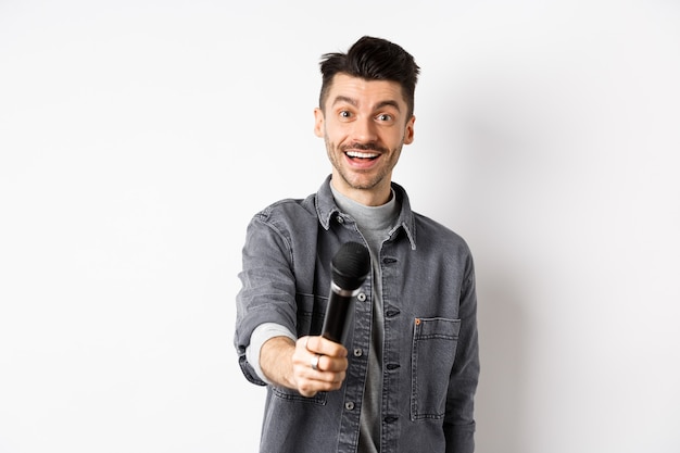 Handsome smiling man stretch out hand with microphone, giving you mic, standing on white background.