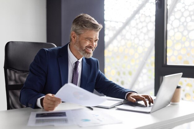 Handsome smiling mature businessman working with documents and laptop in modern office