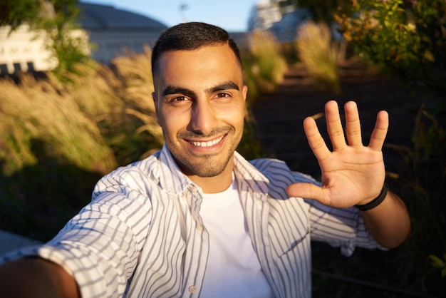 Handsome smiling middle eastern man tourist taking selfie. Influencer waving hand recording video