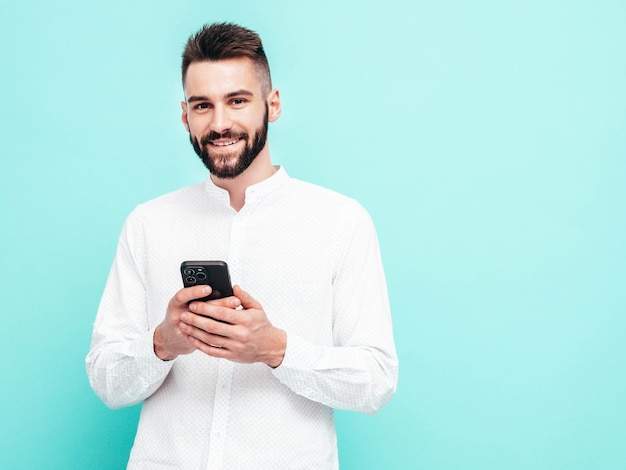 Красивый улыбающийся модель Сексуальный стильный мужчина в рубашке и джинсах Модный хипстерский мужчина позирует у синей стены в студии Держа смартфон Глядя на экран мобильного телефона Используя приложения
