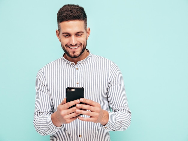 Handsome smiling modelSexy stylish man dressed in shirt and jeans Fashion hipster male posing near blue wall in studio In sunglasses Holding smartphone Looking at cellphone screen Using apps