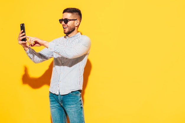 Handsome smiling modelSexy stylish man dressed in shirt and jeans Fashion hipster male posing near yellow wall in studio Holding smartphone Looking at cellphone screen Using apps Holding phone