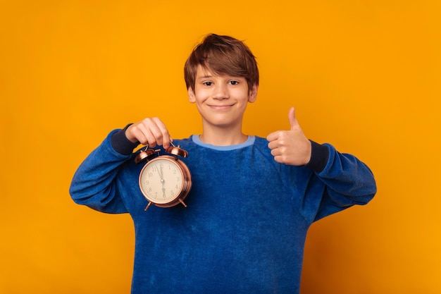 Handsome smiling teen boy is showing thumb up while holding an alarm clock