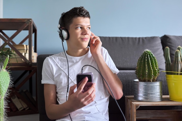 Handsome smiling teenager boy in headphones with smartphone sitting at home, listening music audio blog book, resting, playing, studies. Education leisure lifestyle teens concept