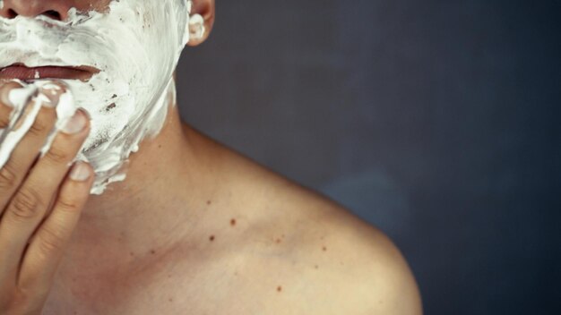 Handsome smiling unshaven young man applying moisturizing shaving foam on face and looking at camera Portrait of happy man performing daily selfcare beauty routine indoors