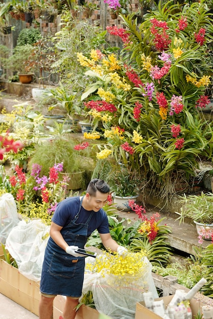 Красивый улыбающийся молодой азиатский работник цветочного питомника с цифровым планшетом проверяет растения и цветы, упакованные и подготовленные для доставки клиентам