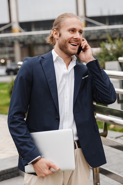 Handsome smiling young bearded businessman standing outdoors in the street, talking on mobile phone carrying laptop