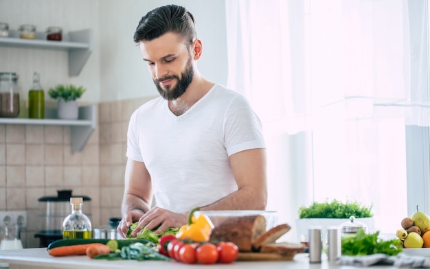 Handsome smiling young bearded man cooks fresh healthy vegan salad for his family and having fun in the big light kitchen