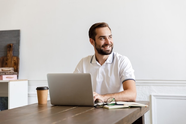 Handsome smiling young bearded man sitting at the table at home, using laptop computer, looking away