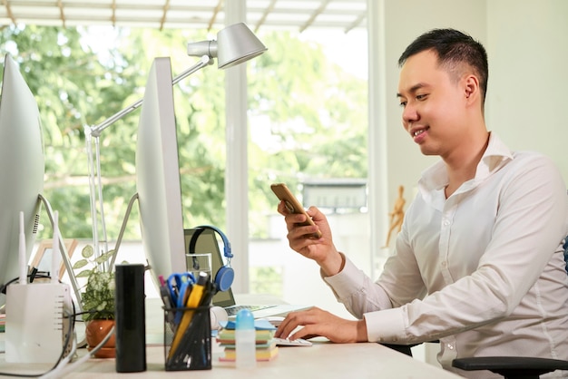 Handsome smiling young businessman sitting at office table and answering text messages on his smartphone