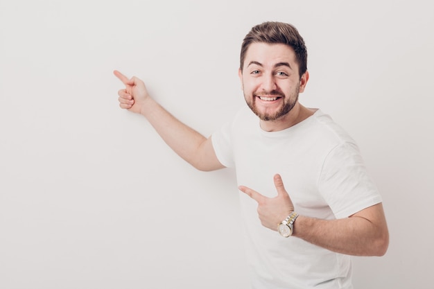 Bell'uomo sorridente con la barba che indica lo spazio della copia. giovane in t-shirt che guarda l'obbiettivo e che punta contro il muro bianco. luce soffusa