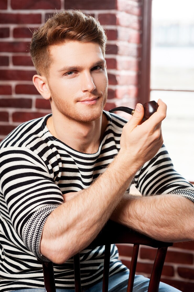 Photo handsome smoking pipe. cheerful young man in striped shirt holding pipe and looking at camera while sitting on the chair
