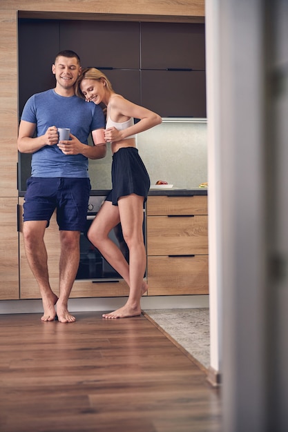 Handsome sporty man and cute blond woman staying in kitchen while drinking tea and talking