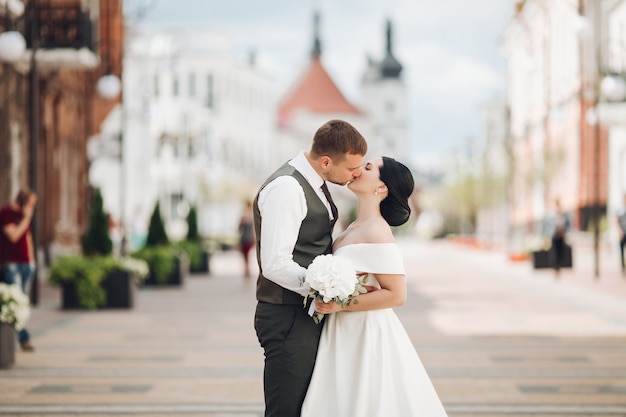 Handsome strong groom kisses with his young pretty bride in white dress with a bouquet of white flowers