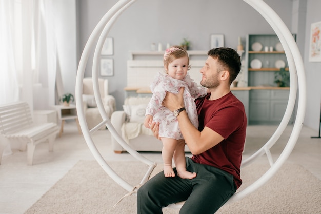 A handsome strong man in a bright room with his cute little daughter sits on a round swing