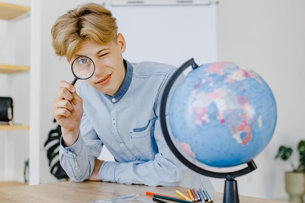 Handsome student is sitting at a desk with magnifying glass in hand Male is watching to the globe in class