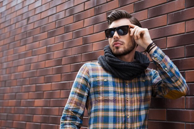 Photo handsome and in style. handsome young man adjusting his sunglasses and looking away while standing against brick wall