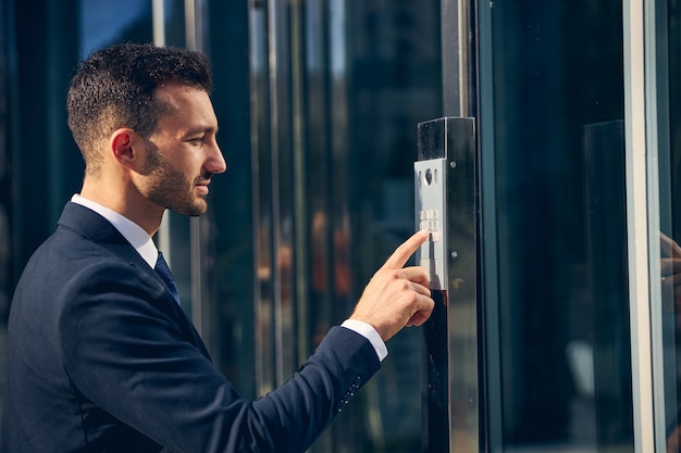 Handsome stylish businessman staying outside in formal clothes while pressing button on building with finger