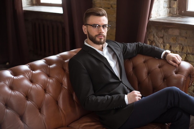 Handsome stylish confident bearded businessman in a smart suit and glasses sitting on a sofa in the office