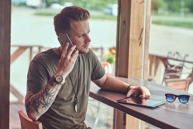 Handsome stylish hipster sits at a table in a roadside cafe, talking on the smartphone.