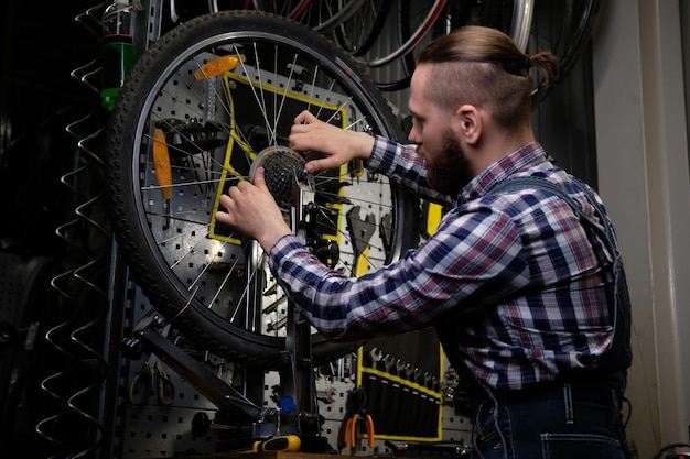 フランネルシャツとジーンズのつなぎ服を着て、修理店で自転車のホイールを操作しているハンサムなスタイリッシュな男性。