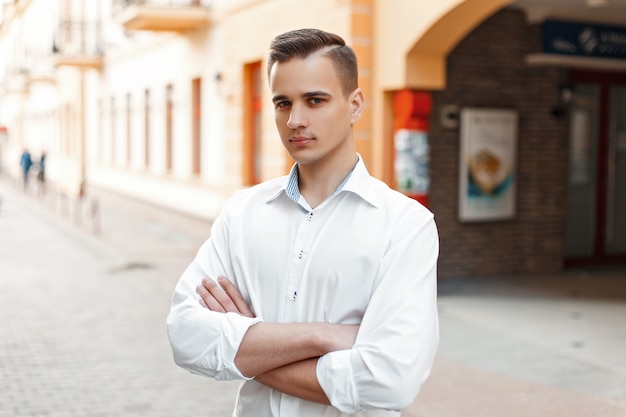 Handsome stylish man with a haircut in a white shirt crossed his arms in the city