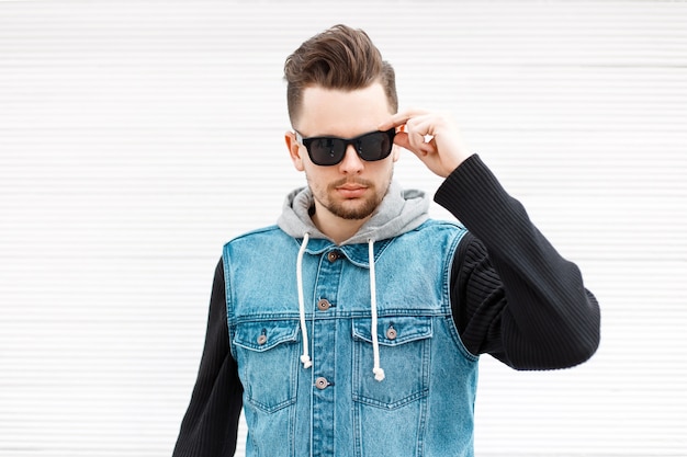 Handsome stylish man with sunglasses in a jeans jacket near a vintage white wooden wall